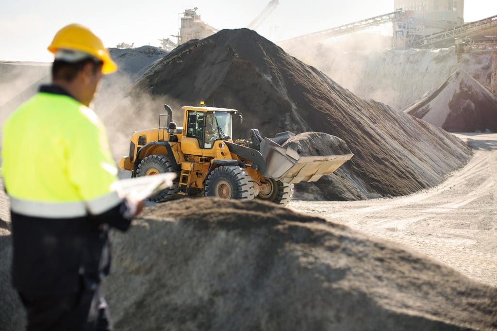 Worker in high-visibility clothing and hard hat holding a clipboard and observing operations at a stone quarry with a front-end loader and large rock piles in the background